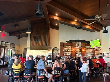 Ranch Bar & Grill Bar Photo Guests seated around the bar at The Ranch Bar & Grill, with bartenders, shelves of drinks, and a TV showing a football game